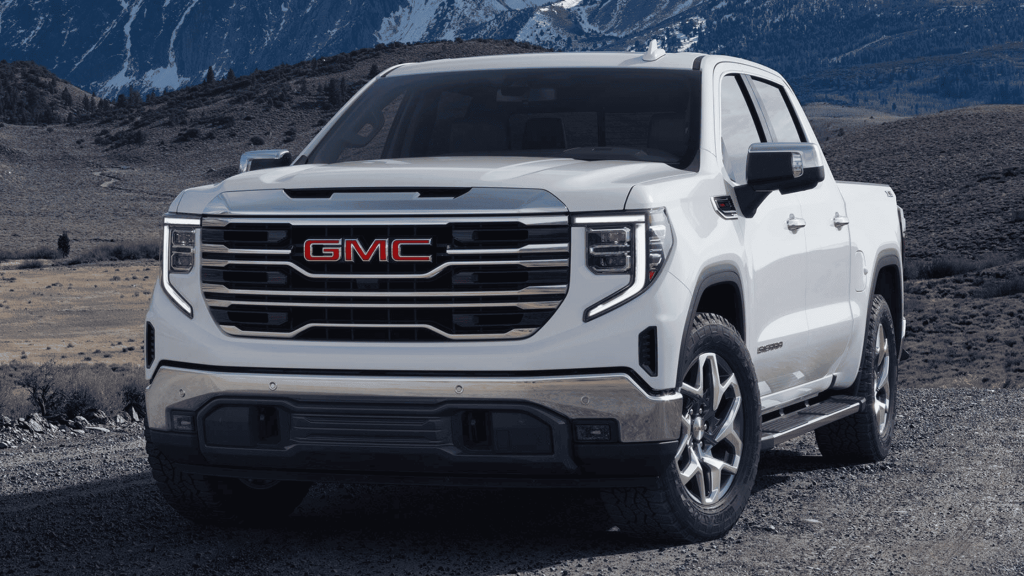 Front view of 2025 GMC Sierra 1500 in white parked on gravel with snow-capped mountains behind