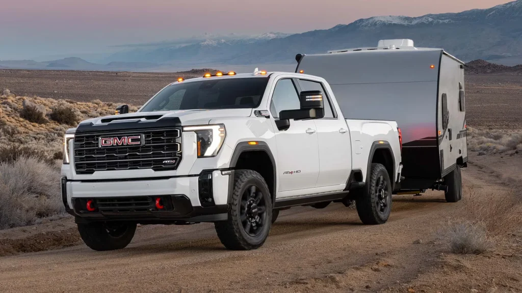 2025 GMC Sierra HD AT4 towing a travel trailer on a rugged desert trail at sunset.