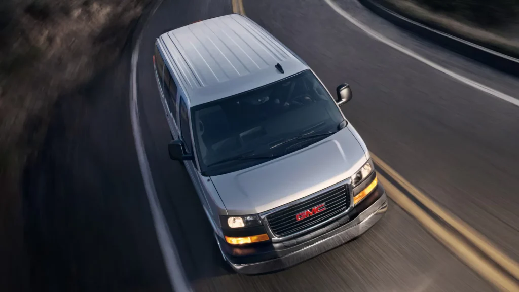 Overhead front view of a silver 2025 GMC Savana Passenger Van driving on a winding highway.