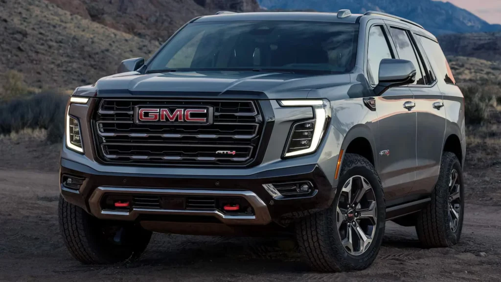 Close-up front view of a gray 2025 GMC Yukon XL AT4 parked on rugged terrain with LED lighting and red tow hooks.