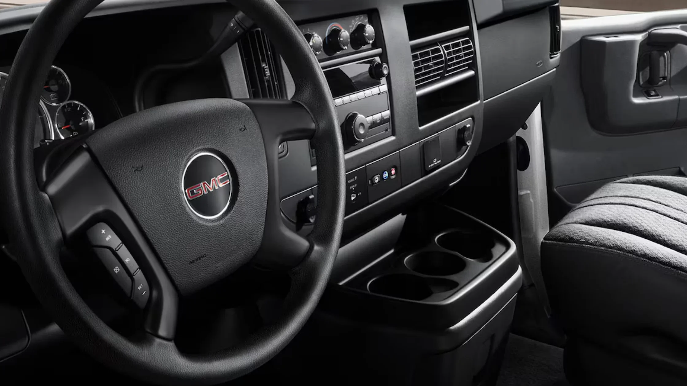 Interior front cabin of the 2025 GMC Savana with cloth bench seat, basic control panel, and traditional black steering wheel.