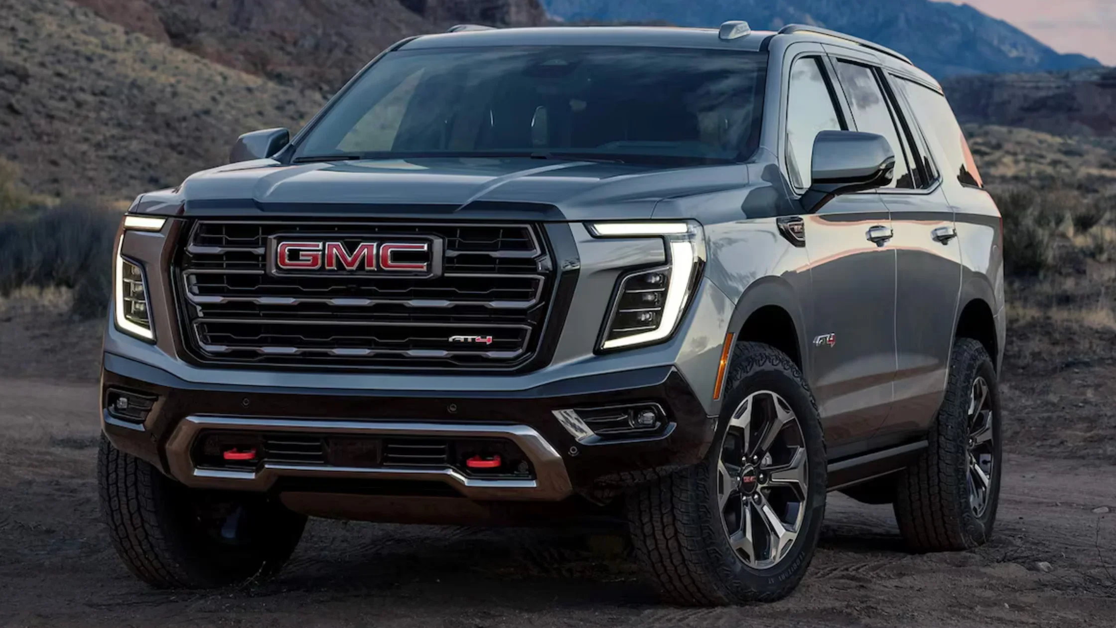 Front view of a 2025 GMC Yukon XL AT4 in dark metallic paint, featuring C-shaped LED headlights, red tow hooks, and rugged off-road styling parked in a desert setting.