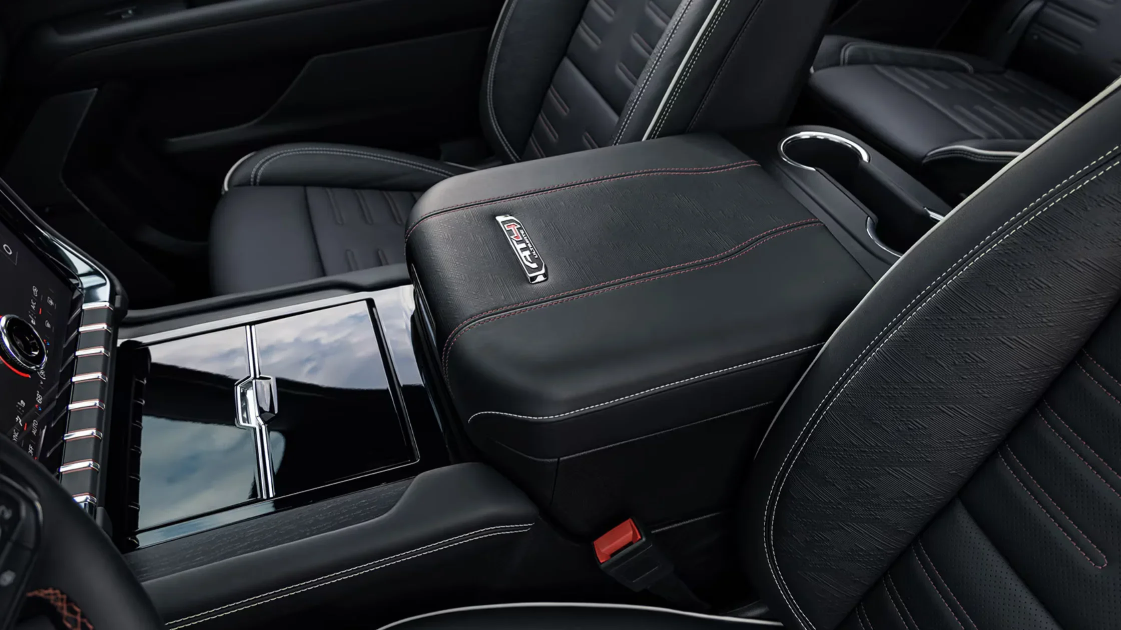 Close-up of the 2025 Yukon XL AT4 center console with black leather armrest, AT4 badge, red stitching, cup holders, and detailed seat texture visible.