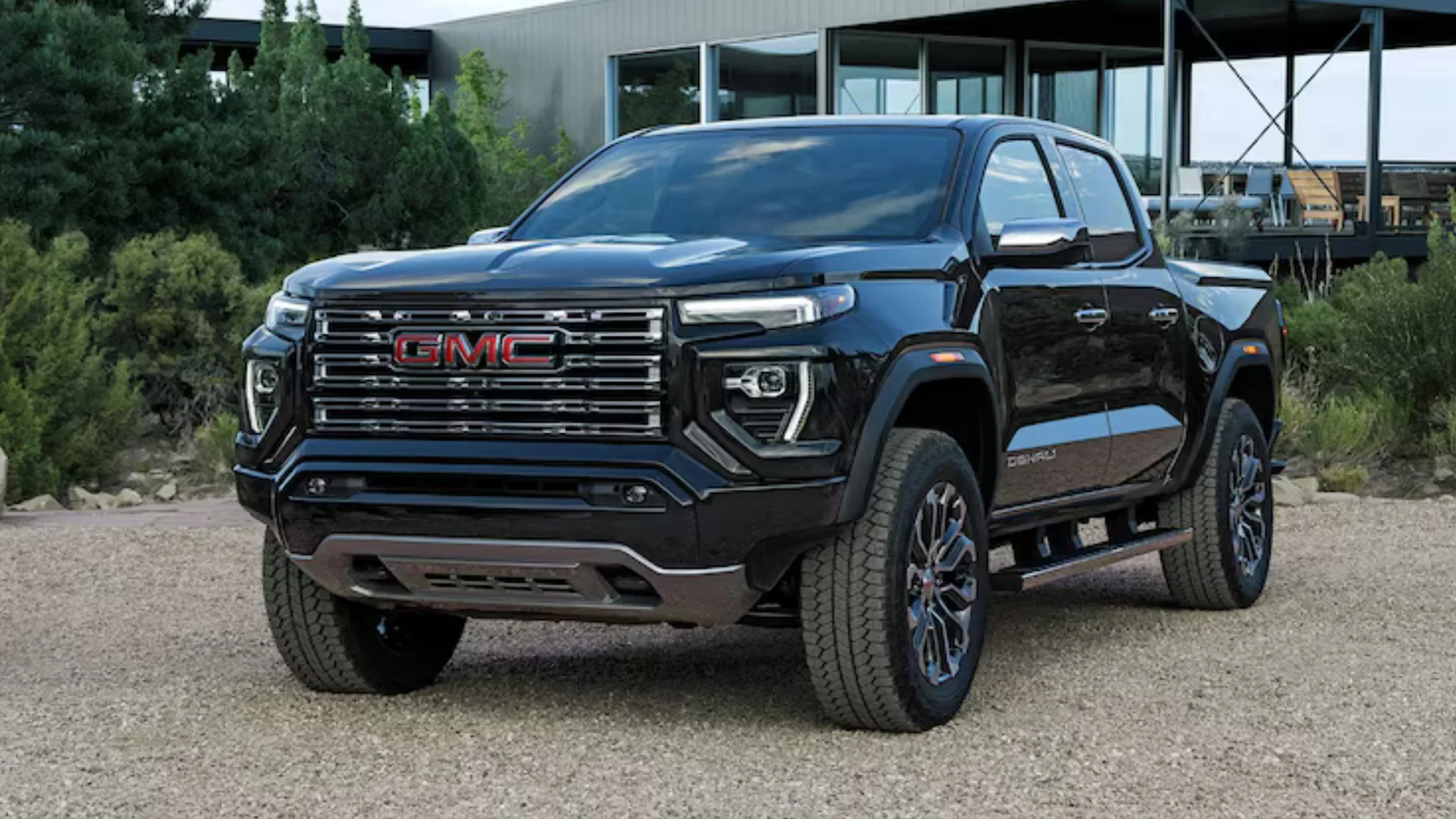 Front angle of 2026 GMC Canyon Denali in black, parked on gravel with modern building in background.